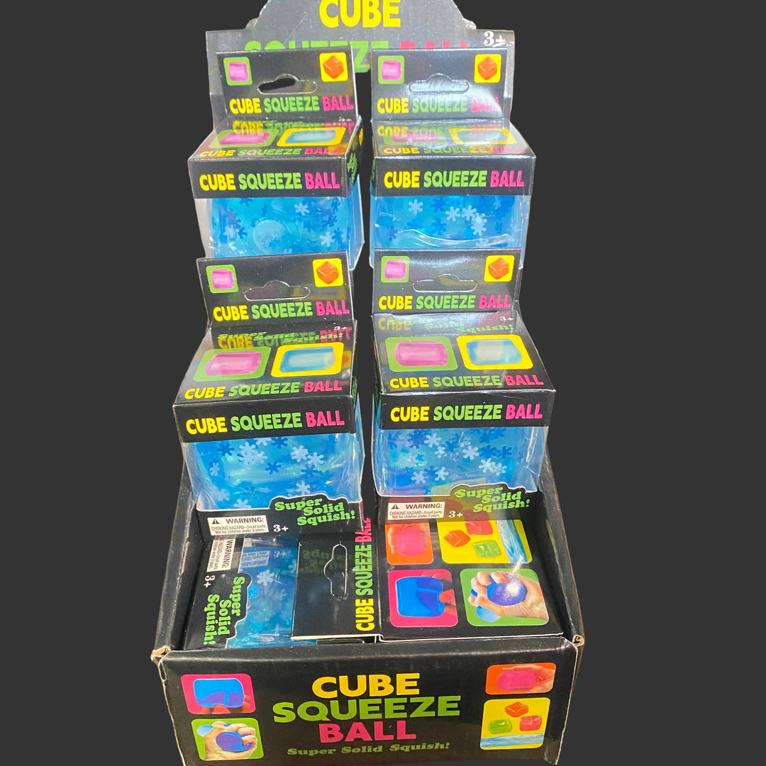 A retail display box filled with multiple packages of Cube Squeeze Ball, a squishy toy. The packages are stacked on a wooden table, with other boxes partially visible in the background.