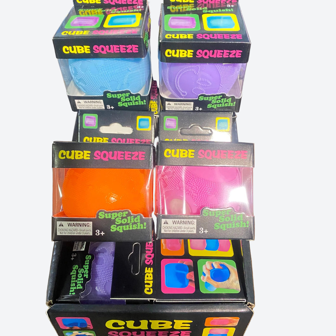 Several boxed Cube Squeeze solid squishy toys arranged on a wooden surface. The packages show different colors including blue, purple, orange, pink, and multicolor square graphics on the front.