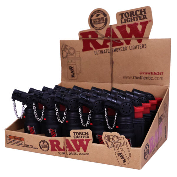 A cardboard retail display box filled with RAW brand torch lighter toys. The display features multiple black lighter-shaped items with silver chains and red accents, arranged inside a brown kraft cardboard box with RAW branding.