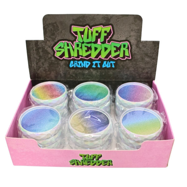 A pink display box of Tuff Shredder 'Grind It Out' makeup shapers, showing six clear plastic cups filled with colorful, iridescent powder that shifts through blue, green, and purple hues.