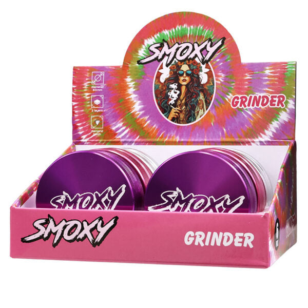 A pink retail display box holding two purple cylindrical metal smokers' grinders. The display features a colorful tie-dye style back panel with the brand name 'Smoxy' and the word 'Grinder'.