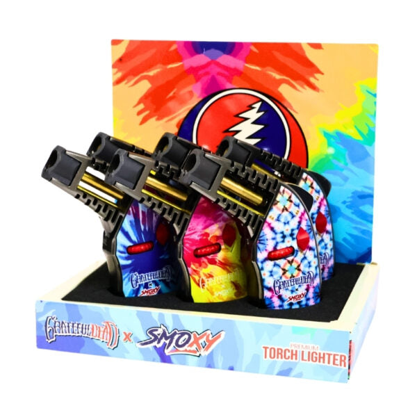 A retail display box containing four Grateful Dead x Smokey Torch Lighters with vibrant tie-dye patterns, set against a plain white background.