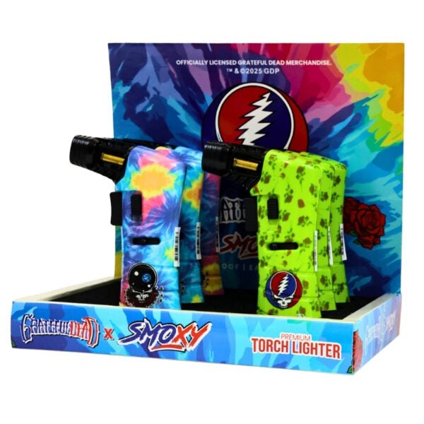 A retail display box containing two Smokey x Grateful Dead themed torch lighters, one with a blue tie-dye pattern and one with a neon green floral pattern, set against a colorful tie-dye backdrop.