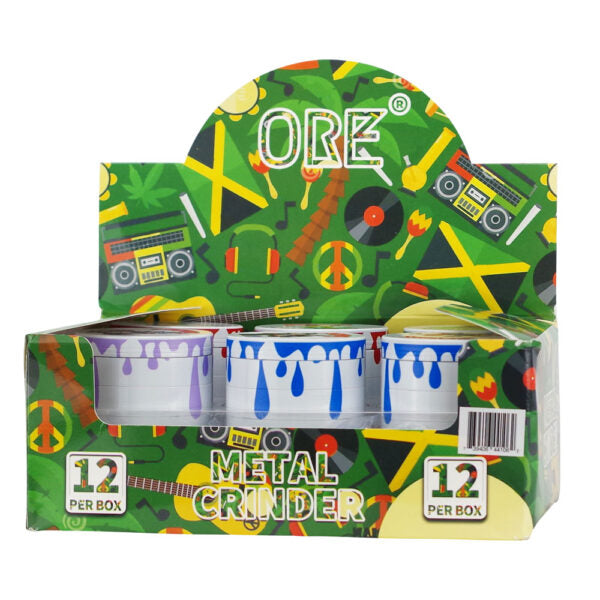 A retail display box of ORE Metal Grinder cups, featuring a green background with a retro 1970s/80s pattern of boomboxes, peace signs, and cassette tapes. The display holds three metal grinder cups with blue, purple, and white paint-drip designs.