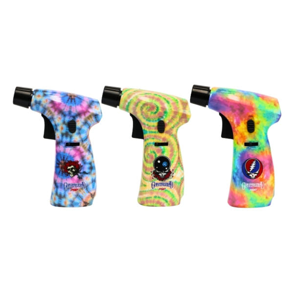 Three handheld torch lighters with colorful tie-dye patterns are shown side by side on a plain white background. Each lighter has a black nozzle and trigger, and the handles feature different vibrant tie-dye designs with Grinch-themed graphics.