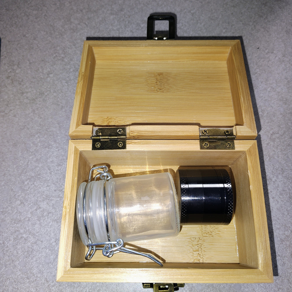 Open wooden storage box containing a clear glass jar with a metal latch and a black metal herb grinder.